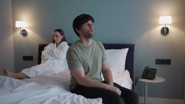 Young angry sad annoyed disappointed swearing screaming couple girl and guy are sitting on the bed indoors in the bedroom at home, spending time in the room