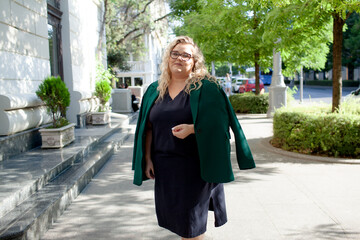 Portrait of a beautiful woman walks in the street of the city. Plus size woman dressed fashion outfit,  green jacket, glasses  and blue dress, posing outdoot