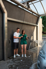 Young pretty caucasian couple holding a water gun on an automatic car wash spraying their car laughing and having fun together