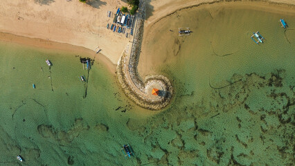 Aerial view of the coastline of Sanur beach in Bali. Gazebo on the curly embankment. Multicolored fishermen's boats are parked near the shore. Beautiful coastline with flat turquoise water.