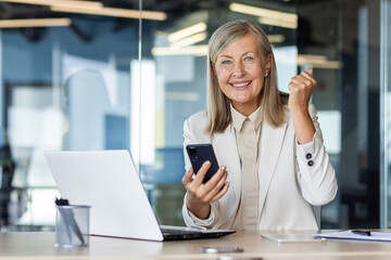 Obraz premium Portrait of a senior gray-haired businesswoman working in the office at a laptop, holding a phone in her hands, happy and smiling at the camera, showing a victory gesture with her hand.