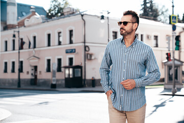 Portrait of handsome confident stylish hipster lambersexual model. Sexy modern man dressed in blue shirt and trousers. Fashion male posing on street background in Europe city at sunset. In sunglasses