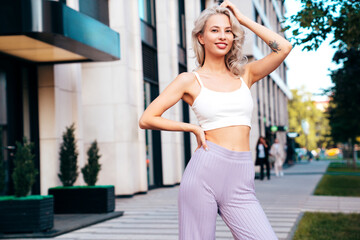 Fototapeta na wymiar Young beautiful smiling hipster woman in trendy summer white top and violet pants clothes. Carefree model posing in the street at sunset. Positive blond female. Cheerful and happy
