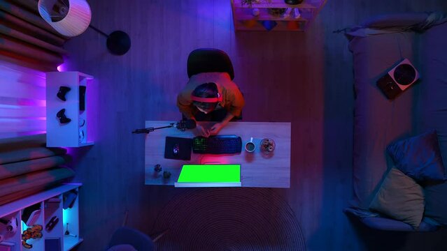 Top view of man in headphones streaming playing game on PC with Chroma key green screen, talking drinking coffee, advertising area workspace mockup.