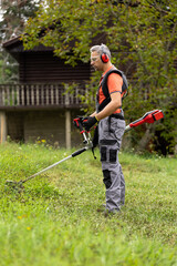 Young adult man in protective clothes mowing green grass with electrical lawn trimmer at garden. Side view.