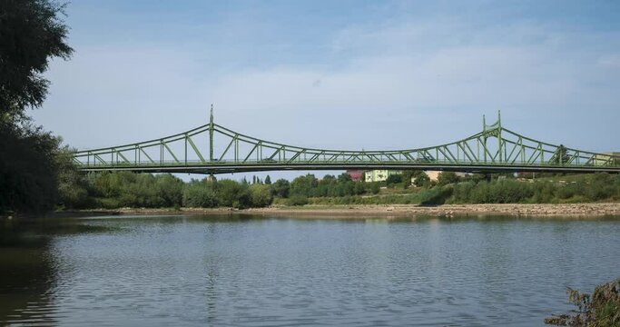 Time lapse of traffic driving on an old steel bridge over a narrow European river. Summer day, thin clouds, green trees, no people