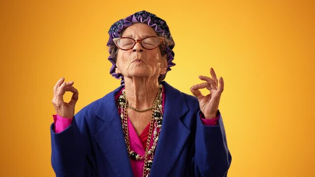 Funny crazy grandmother senior woman saying OM, 80s, meditating doing yoga exercise isolated on yellow background. Concept of youthful old female.