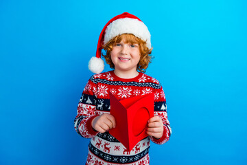 Photo of cheerful charming little boy dressed ugly pullover x-mas hat preparing gift card isolated blue color background