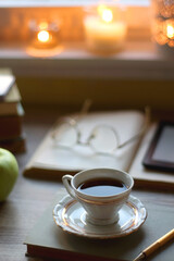 Vintage books, e-reader, pen, reading glasses, phone, cup of tea or coffee, bowl of cookies, green apple and lit candles on the table. Hygge at home, studying atmosphere. Selective focus.