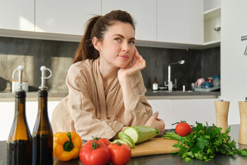 Portrait of pretty woman in bathrobe, standing in kitchen with thoughtful face, posing near vegetables and bottle of oil, thinking what food to prepare, making salad, eating healthy - Powered by Adobe
