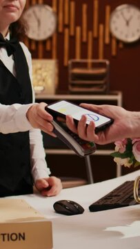 Vertical Video Elderly man makes nfc transaction with mobile phone at pos terminal, paying for accommodation at five star resort. Tourist arriving at front desk doing check in for holiday. Close up