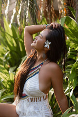 Side view of young smiling gorgeous Asian woman holding long dark hair sitting near palm trees plants, looking up.