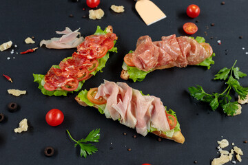 Bruschetta with salami, chorizo and prosciutto on a black background with ingredients for cooking.