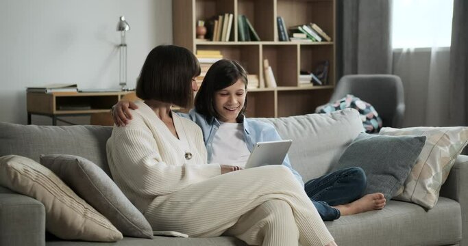 Contented mother and son sit on the couch with a digital tablet, sharing laughter and joy. Their happiness creates a heartwarming and cheerful atmosphere in the room.