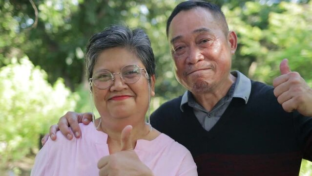 Portrait of an elderly Asian couple smiling happily in the garden, both giving thumbs up. Living a happy retirement life. health care