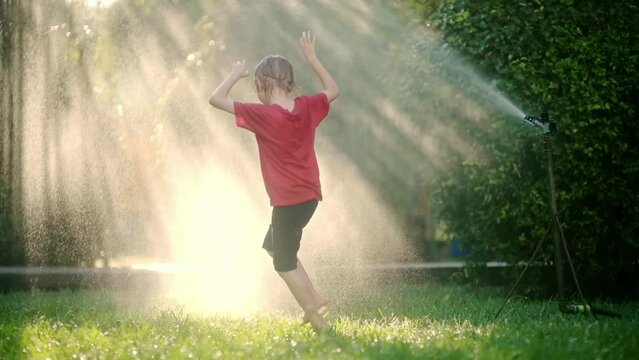Slow motion video of funny little boy playing with garden sprinkler in sunny backyard.Elementary school child laughing, jumping and having fun with spray of water.Summer outdoors activity for children