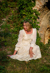 barefoot beautiful blonde woman in a white dress and wreath from green leaves posing in ruins near...