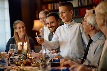 Happy Jewish boy lighting menorah during family meal on Hanukkah.