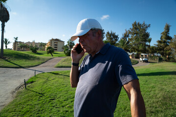 Older golf player talking on a cell phone on the course of a golf club. Sunny day, wearing a white cap