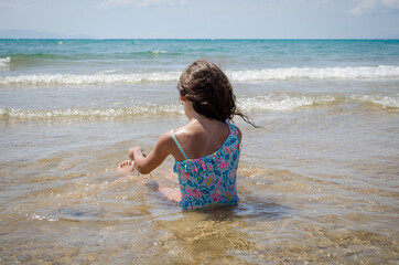 Brunette Girl with Blue Swimsuit Enjoying Shallow Sea at Sandy Beach. Tranquil Coastal Scene in Greece. Horizon and Sky in Background.