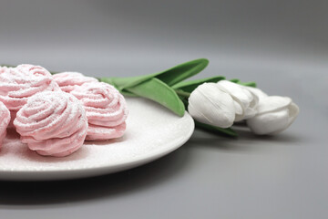 Delicate handmade pink marshmallows on a white plate with white tulips on a gray background.