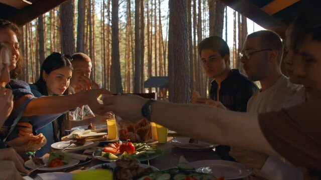 Friends eat in gazebo in nature. Stock footage. Relaxing with friends and barbecue in woods on sunny summer day. Friends eat barbecue at table