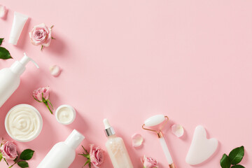 Flat lay composition with cosmetics packages, jars of cream, serum bottles, gua sha, face roller with rose buds and petals on light pink background. Natural beauty products for face skin care concept.