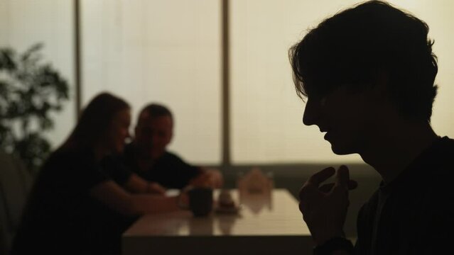Man at the fast food bistro bar eating with friend and talking, girl with man at the background watching smartphone.
