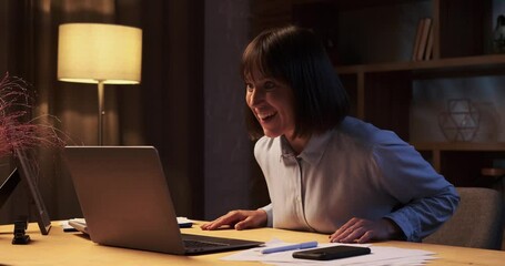 Cheerful Caucasian woman reacts with genuine joy to good news on laptop. Face lights up with a radiant smile and excitement. The room is filled with a sense of accomplishment and positivity