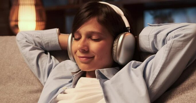 Happy schoolboy sits on the sofa at evening living room, immersed in the melodies streaming through headphones. His face radiates pure joy as he enjoys the music, eyes closed in bliss.