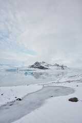 A serene wintry scene with a vast lake and snow-capped mountains. Icebergs float in the lake under a clear blue sky.