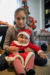 two sisters sitting in front of the Christmas tree, the older one hugs the little one