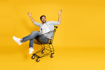 Happy young man sitting inside of shopping trolley isolated on yellow background, Wow and surpise concept