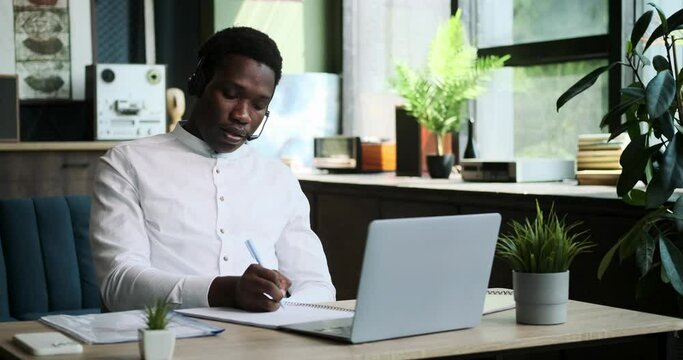 A professional Black manager, equipped with a headset, conducts a business call from the comfort of home. His efficient and focused demeanor reflects his commitment to work responsibilities.