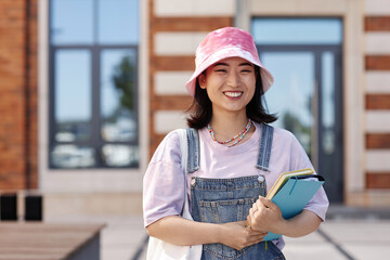 Waist up portrait of fun young Asian woman wearing denim on college campus and smiling at camera,...