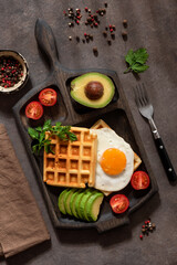 Waffles, fried egg, avocado and tomatoes on a dark background with shadows. Top view, flat lay. Healthy breakfast.