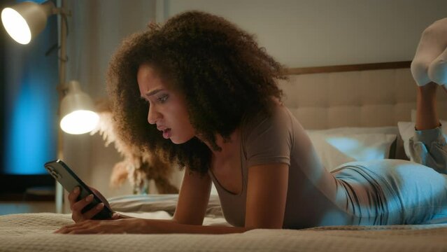 African American young ethnic woman upset worried anxious girl reading bad news on mobile phone lying on bed in night home bedroom sad shocked female using smartphone error notification losing game