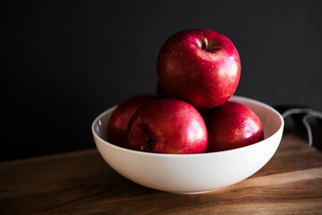 Bright white bowl of apples on wooden cutting board with dark moody background rustic fall autumn feeling
