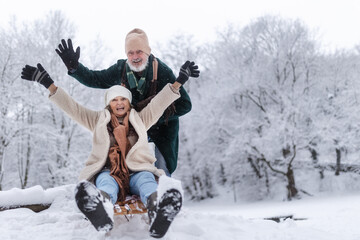 Senior couple having fun during cold winter day, sledding down the hill. Winter vacation in the mountains. Wintry landscape.