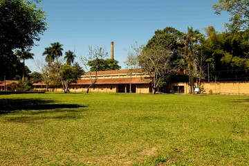 Prédio de fabrica abandonado com chaminé e parede construídas com tijolo a vista cercada com árvores e gramado. 