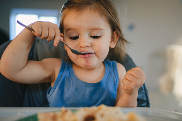 Front view portriat of a child himself eating food at home in the kitchen. The child is sitting in a high chair. The girl eats with a spoon.