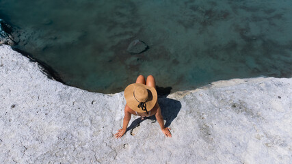 Top view of a woman in a swimsuit with a hat relaxing sitting on the edge of a small natural rock...
