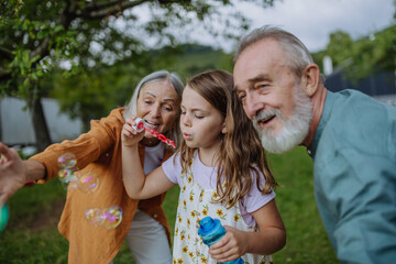 Obraz premium Grandparents and granddaughter are blowing bubbles from a bubble wand, having fun outdoors in the garden during warm autumn day.