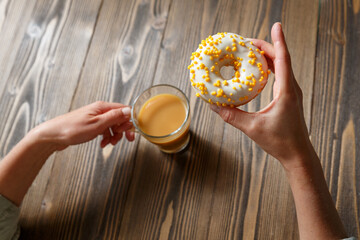 Female hands holding cup of coffee and donut in a white glaze with yellow confectionery sprinkles on a wooden background. - 660106001