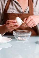 Female hands peeling off boiled egg shell on a kithcen table over glass plate.