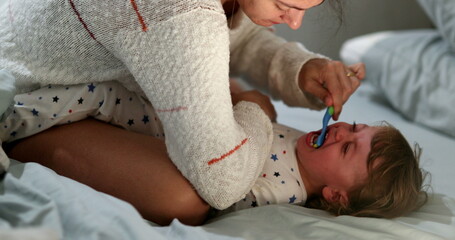 Candid mother brushing crying baby teeh. Upset infant boy not wanting to brush teeth - obrazy, fototapety, plakaty