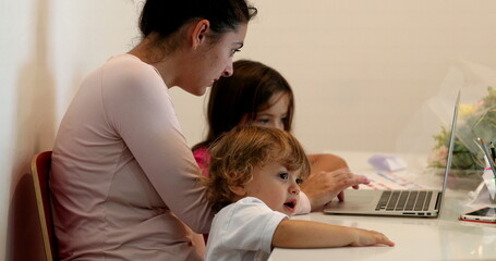 Candid mother multi-tasking at home in front of laptop and parenting kids