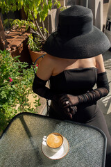 Pregnant beautiful elegant lady in a black dress and a black hat with a large pearl necklace drinks...