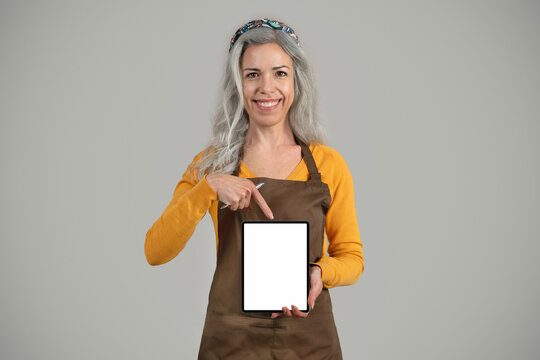 Smiling senior european woman gray hair framer in apron point finger at banner with free space