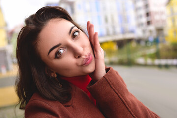 Cute young woman in a brown autumn coat takes a selfie on the street for her boyfriend.